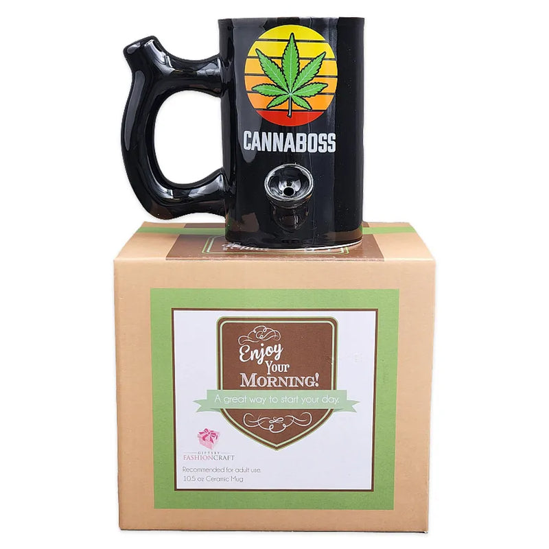 A black ceramic smoking pipe shaped as a mug with a weed leaf decal on top of a sunset background and the word cannaboss underneath on top of its display box.