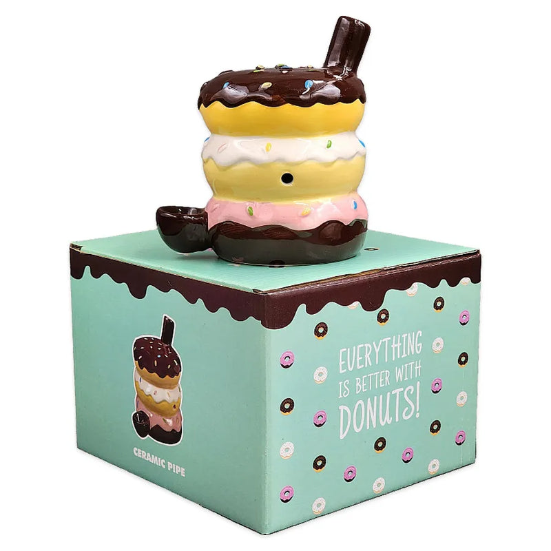 A ceramic smoking pipe in the shape of three donuts stacked on top of eachother. A chocolate covered donut on top, vanilla covered donut in the middle, and a strawberry covered donut on the bottom. On top of its display box.