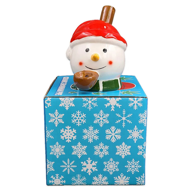 A ceramic smoking pipe that looks like a snowmans head wearing a santa hat and a green scarf on top of its display box.