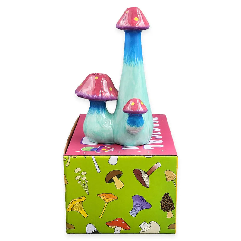 A smoking pipe shaped as 3 mushrooms and made from ceramic. Deep hues of blue and shades of green with a pinkish purple mushroom head that has yellow spots. On top of its display box.